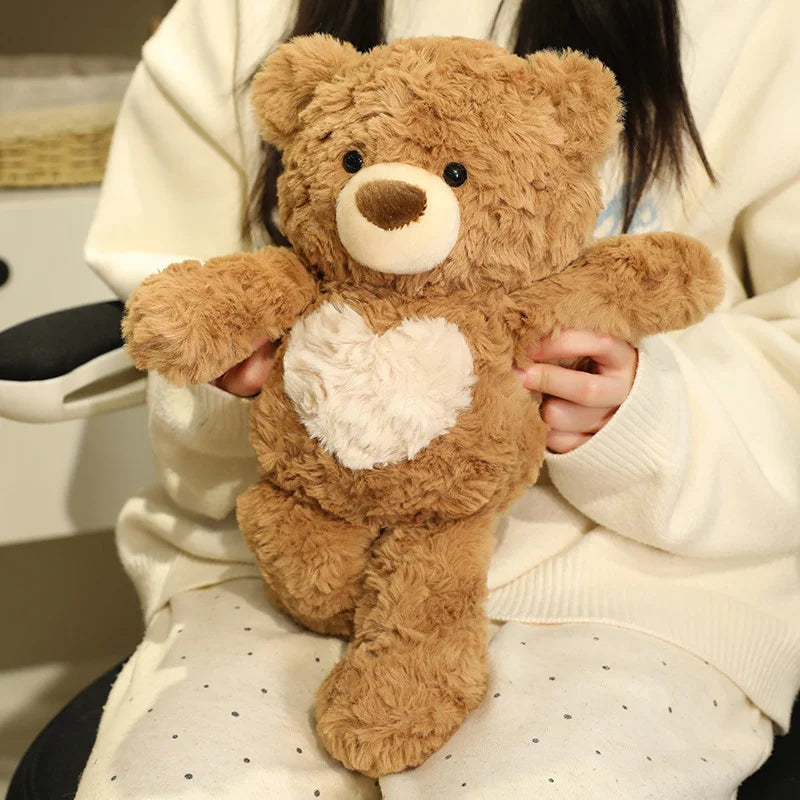 A brown teddy bear with a white heart patch is held by a person wearing a cream-colored sweater.