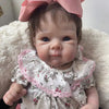 A lifelike 18-inch reborn baby doll with hand-painted details wearing a white floral dress and pink bow.