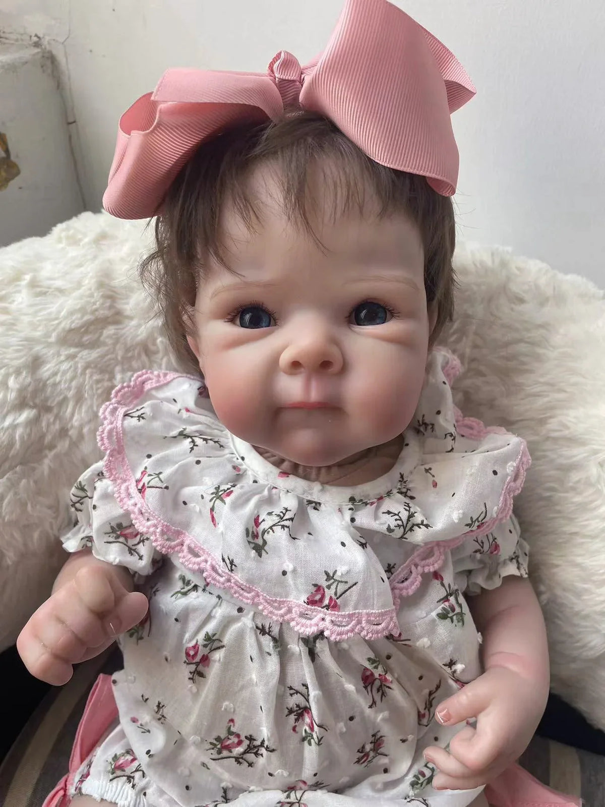 A lifelike 18-inch reborn baby doll with hand-painted details wearing a white floral dress and pink bow.