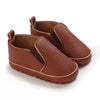 A pair of brown faux leather baby first walker sneakers with a dinosaur print detail.