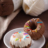 A brown donut-shaped pastry decorated with colorful sprinkles is shown next to a ball of white yarn and a roll of brown yarn.