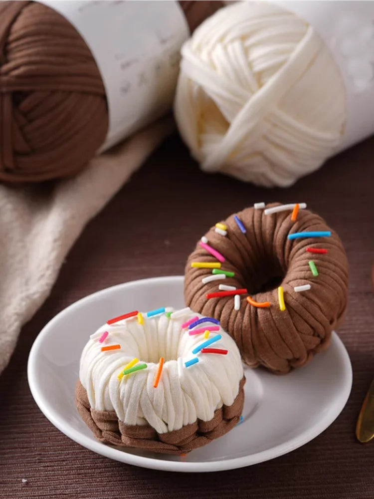 A brown donut-shaped pastry decorated with colorful sprinkles is shown next to a ball of white yarn and a roll of brown yarn.