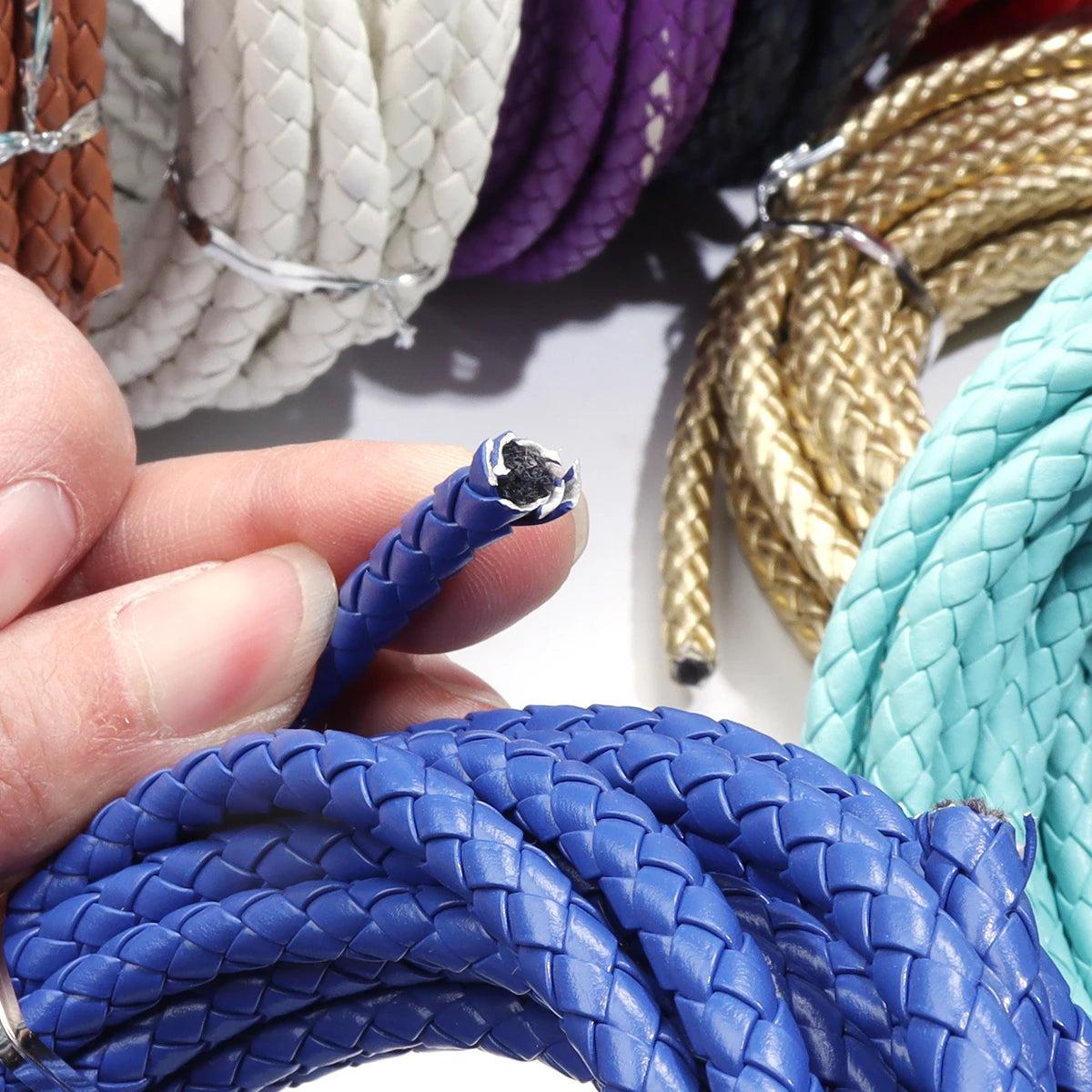 A hand holds a blue round braided leather cord next to various colored cords for jewelry making and crafts.