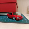Modelo de coche rojo a escala 1/64 posicionado sobre una alfombrilla de rejilla con ruedas y neumáticos.