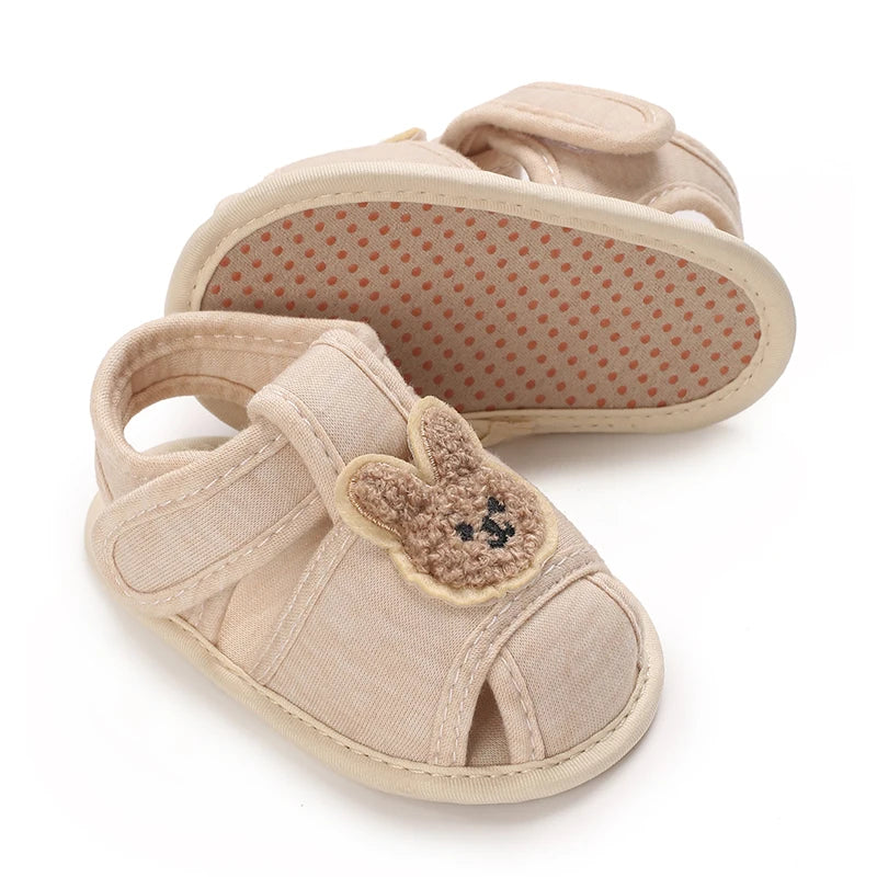 A pair of beige soft sole sandals for a baby, featuring a bear-shaped embellishment.