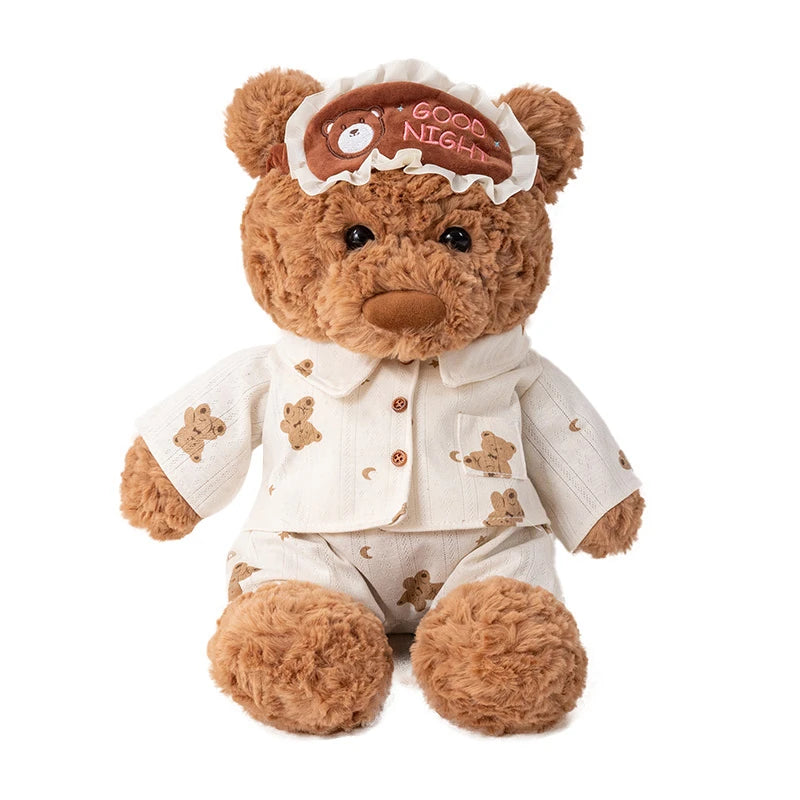 A brown teddy bear plush toy wearing a light beige outfit and an eye mask with the words GOOD NIGHT.