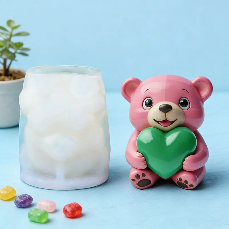 A pink silicone bear mold shaped like a hug with a green heart, placed next to a translucent white candle.