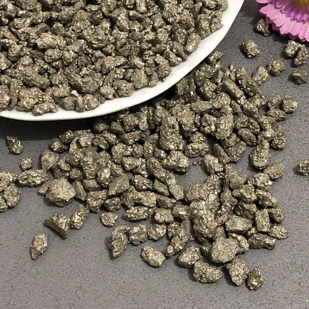 A pile of metallic gold crystal chips, including chalcopyrite and pyrite mineral decor, is shown on a gray surface.