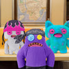 A collection of three plush toys resembling ugly monsters with human-like teeth, featuring purple, gray, and bright blue fur.