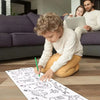 A young boy is drawing on white sticky coloring paper with a green pencil on a wooden floor.