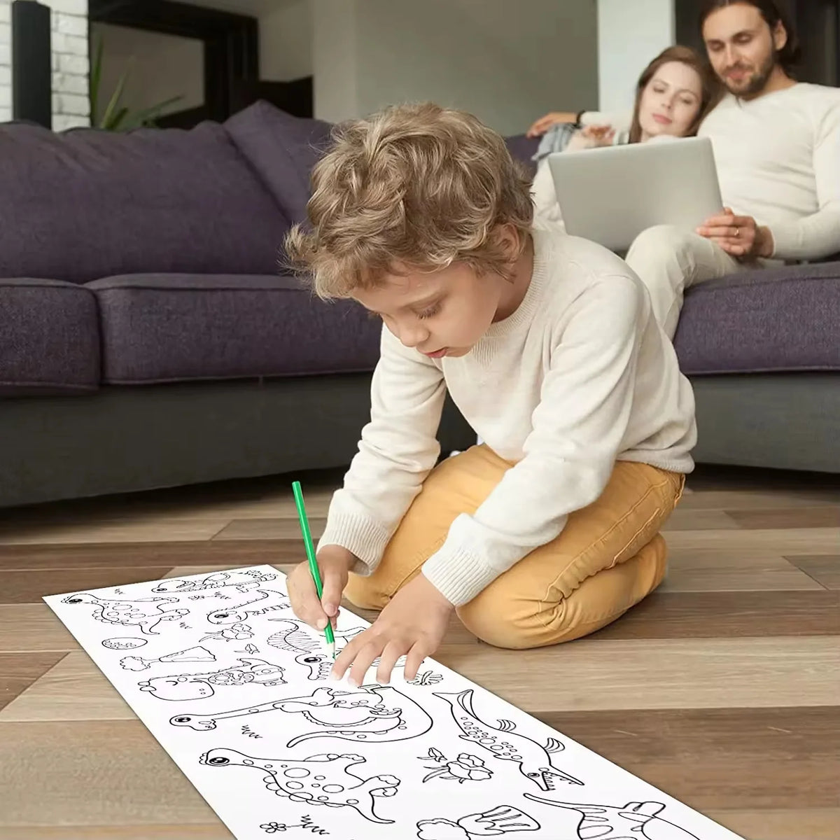 A young boy is drawing on white sticky coloring paper with a green pencil on a wooden floor.