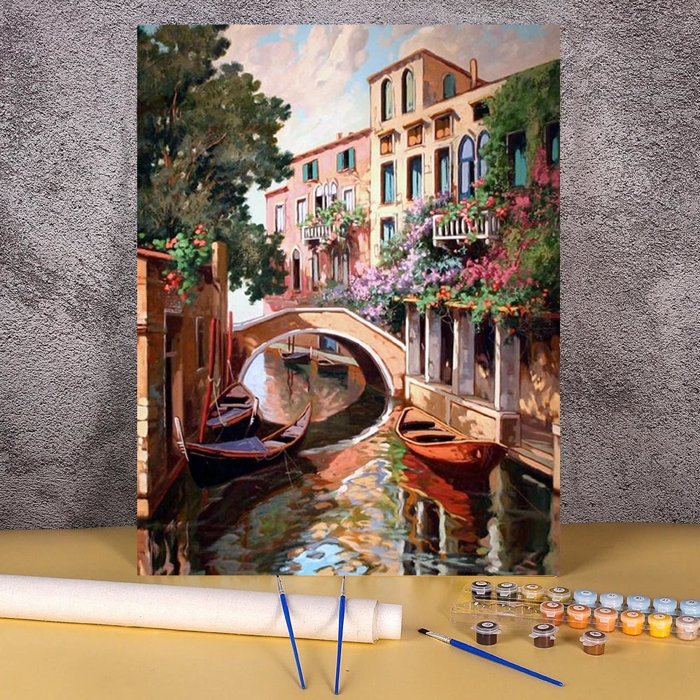 A painting by numbers kit depicting a landscape of a wooden house and flower field over a canal.