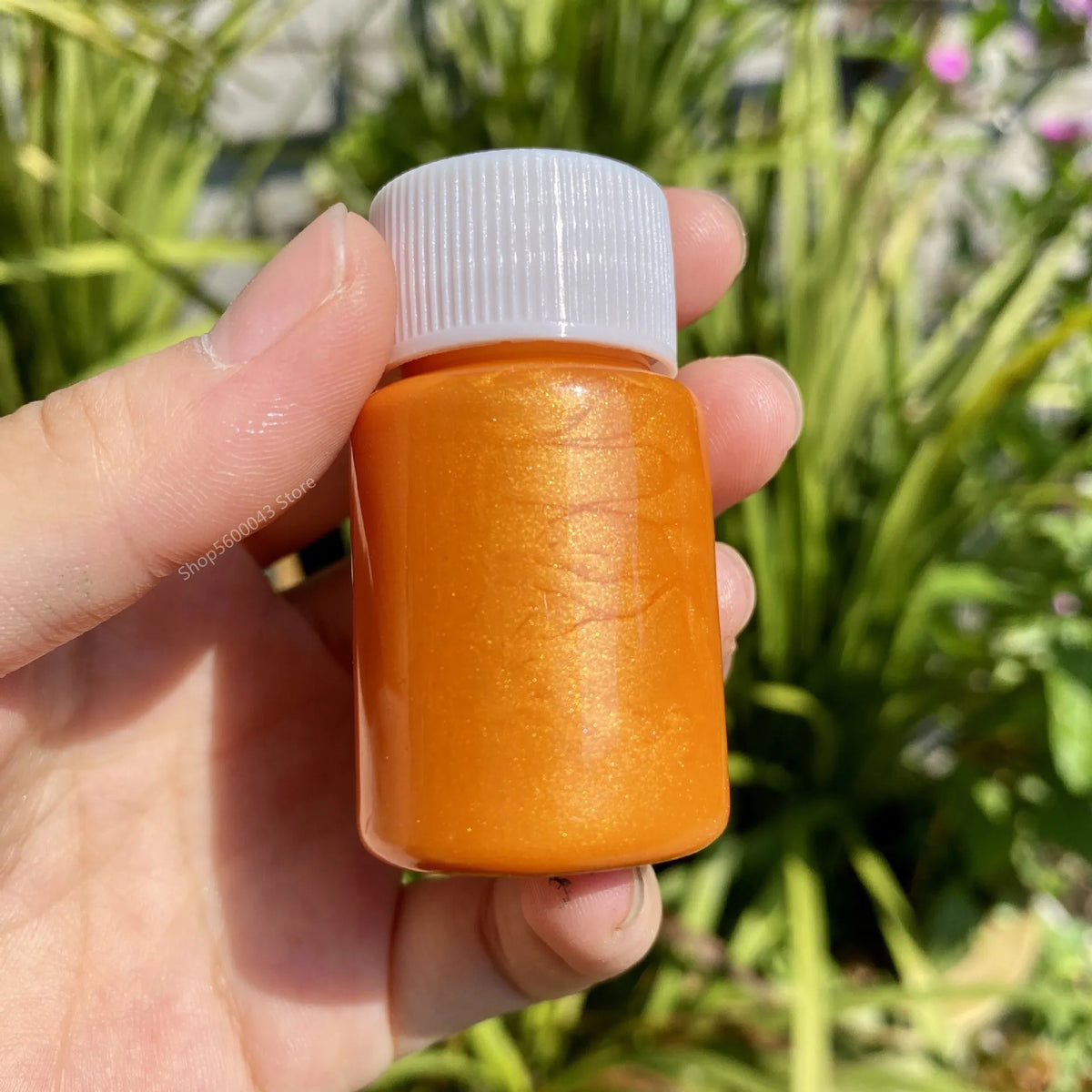 A hand holds an orange metallic gold acrylic paint bottle with a white cap against a blurred green outdoor background.
