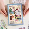 A colorful sticker book featuring retro themed illustrations and a magic display window is held by hands.