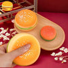 A hand holds an orange squishy hamburger stress ball with white sprinkles on a wooden surface.