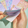 A hand holds a stack of colorful fantasy aesthetic stickers and a pink card featuring more designs.