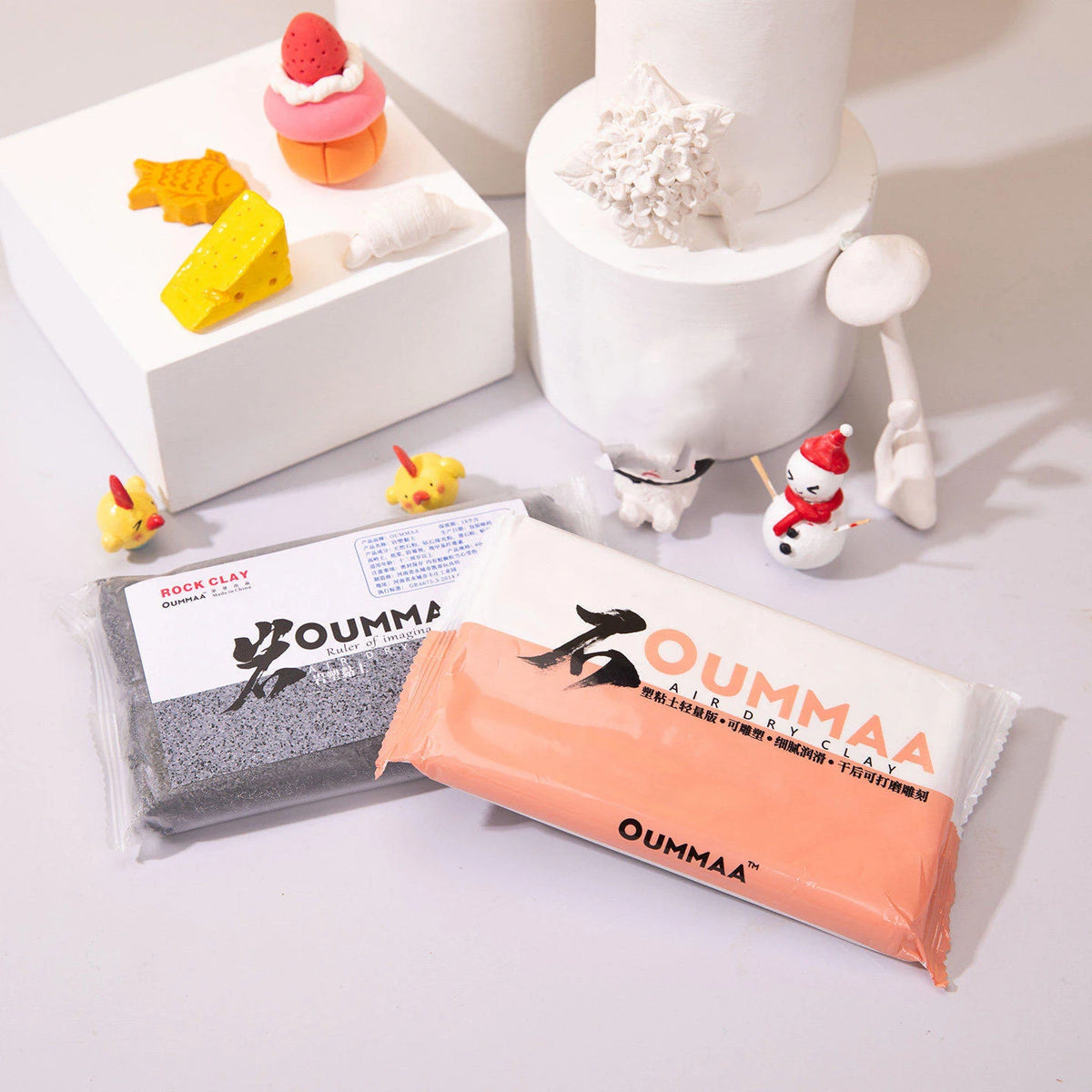 A white cake decorated with orange and yellow clay figures, next to a package of OUMMAA air-dry stone effect modeling clay.