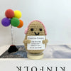 A handwoven crochet potato ornament with pink accents sits on a wooden base next to colorful pom-poms.