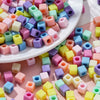 A close-up view of numerous square acrylic spacer beads in various pastel colors scattered on a pink surface.