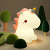 A white unicorn silicone LED night light with a pink horn and rainbow tip sits among various green and dark colored silicone 
