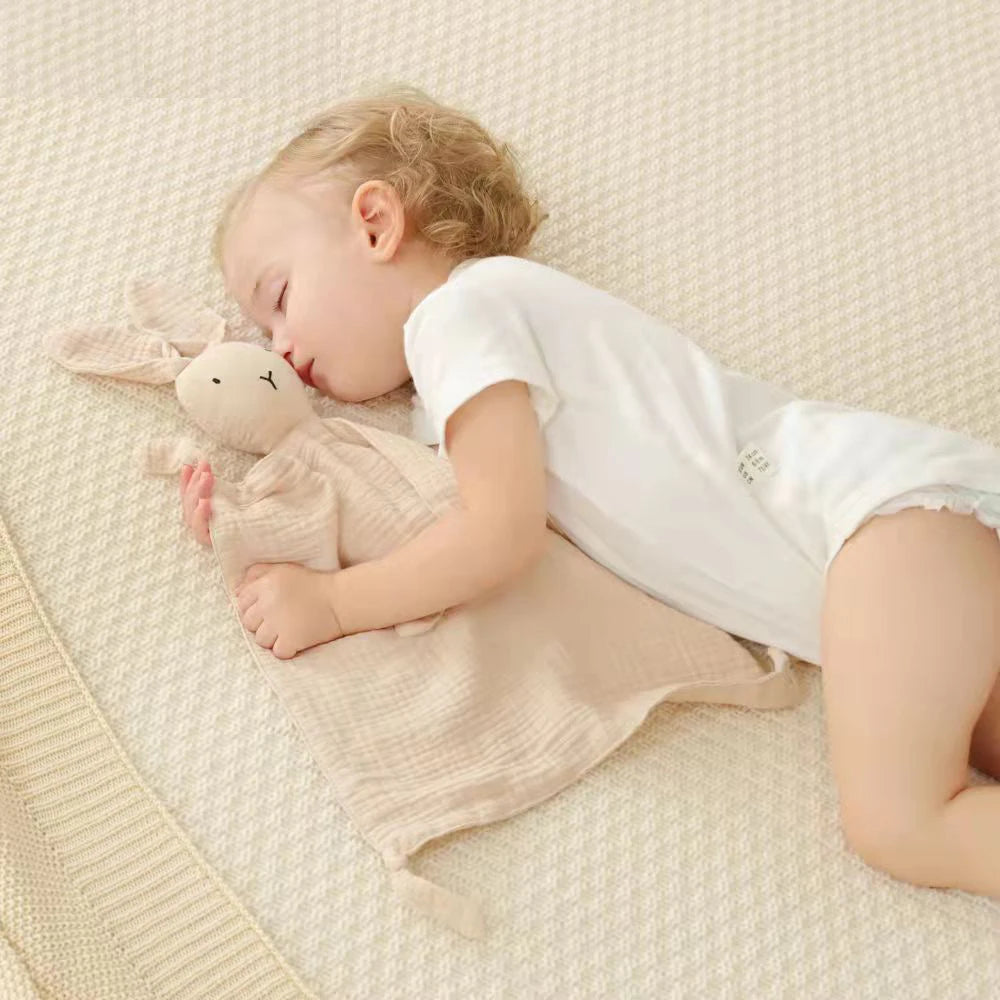A baby is sleeping peacefully on a light beige fabric with a stuffed animal, using a soft cotton muslin comforter blanket.