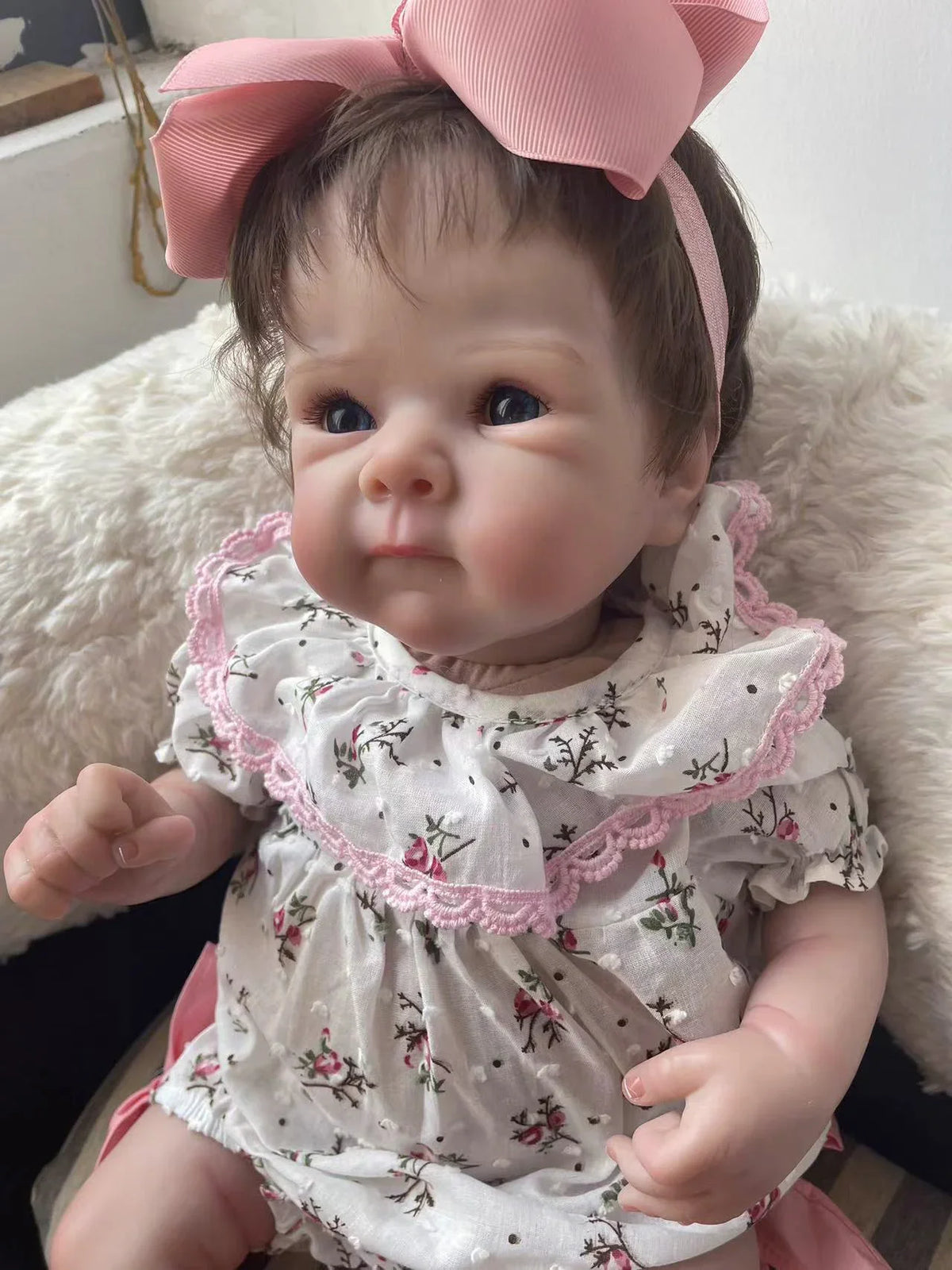 A lifelike 18-inch reborn baby doll with hand-painted details wearing a pink floral dress and bow.