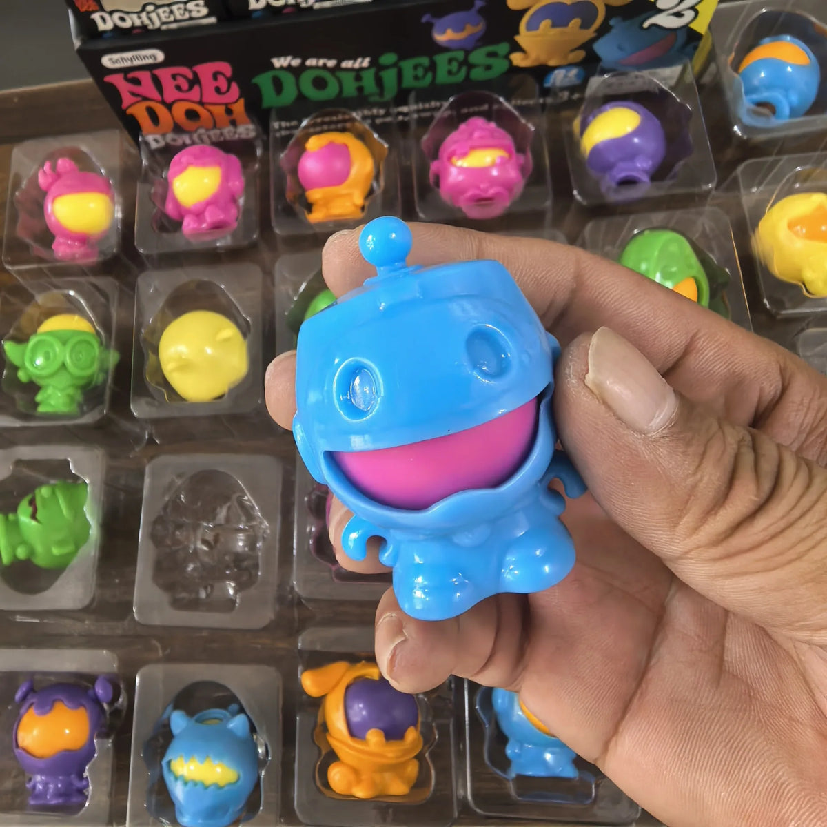 A hand holds a blue squishy fidget toy from the Nee DohJees line, surrounded by various colorful squishies.