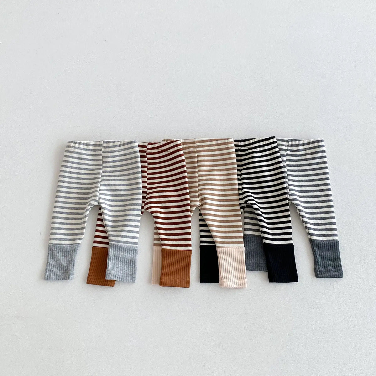 A row of baby leggings featuring various striped and solid color designs in gray, brown, beige, black, and white.