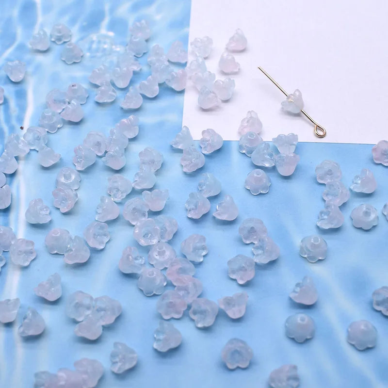 A collection of small, translucent pink and blue glass beads resembling lily of the valley flowers scattered on a light blue 