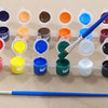 A row of twelve small pots of acrylic paint in various colors arranged on a white surface with a paintbrush resting below.