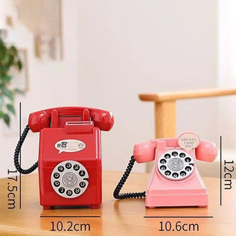 A red vintage rotary telephone piggy bank with a coiled cord and a pink version next to it on a wooden surface.