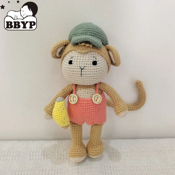 A handmade crochet monkey plush toy with a green cap and pink overalls sitting on a white surface.