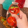 A hand holds a clear bottle shaped like a strawberry with a green top, part of a fruit bottle stress ball set.