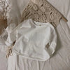 A white kids long sleeve cotton tee is laid on a white sheet next to a beige knitted item.