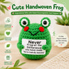 A handmade crochet frog doll in green with red eyes and a motivational card on a wooden surface.