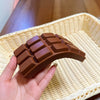 A hand holds a realistic brown chocolate squishy toy resting on a woven tray.