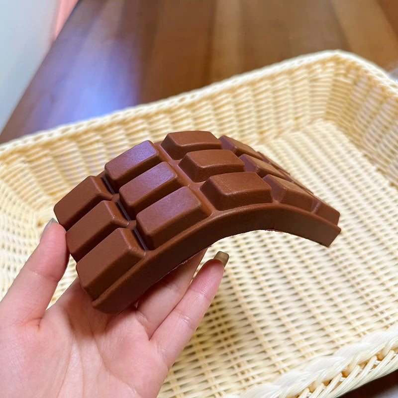 A hand holds a realistic brown chocolate squishy toy resting on a woven tray.
