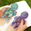 A hand holds a translucent purple squishy baby stress relief toy with glittery accents on a white notebook.