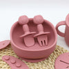 A pink silicone baby tableware set featuring a spoon and fork with suction cups resting on a woven mat.