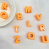 Orangefarbene Acryl-Buchstabenperlen, die ROMV L ZOUR buchstabieren, verstreut auf einer weißen Fläche neben einer kleinen Sc