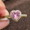 A close-up of a gold heart-shaped ring featuring a large pink gemstone surrounded by small clear stones.