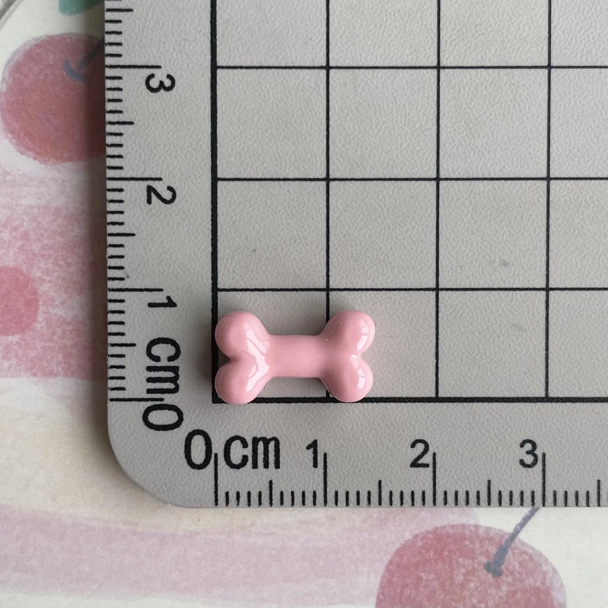 A small pink resin bone charm is placed on a ruler marked in centimeters and inches.