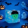 A clear blue ice cube shaped like a ghost is resting on a dark surface with Halloween-themed decorations in the background.
