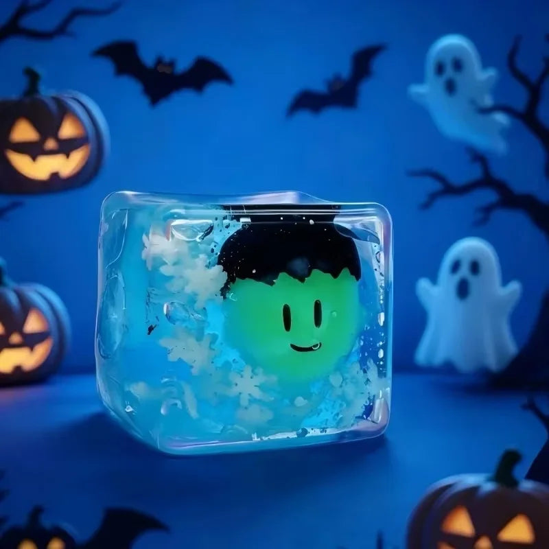 A clear blue ice cube shaped like a ghost is resting on a dark surface with Halloween-themed decorations in the background.
