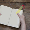 A hand is holding an open book and placing a yellow bookmark inside, next to a small red cartoon desk lamp.