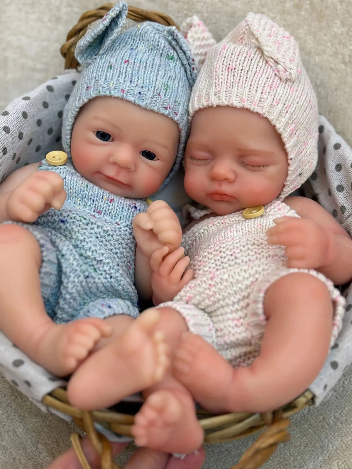 Two newborn silicone reborn dolls with knitted hats are placed in a woven basket.