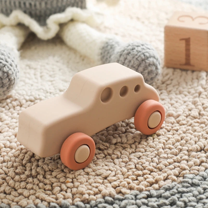A beige silicone baby teether toy shaped like a cartoon car with round orange wheels resting on a knitted fabric.
