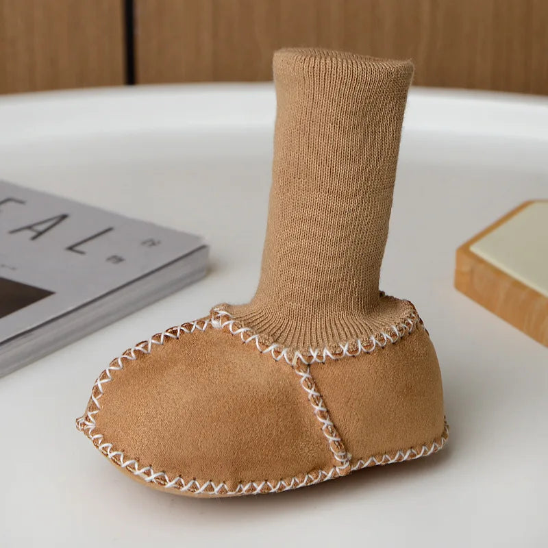 A pair of tan suede baby booties with a ribbed cuff and white stitching, resting on a white surface.