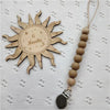 A wooden sun-shaped pacifier clip with six months engraved on it, attached to a string of natural beads.