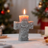 A white floral cross candle with a lit wick sits on a table in a softly lit holiday setting.